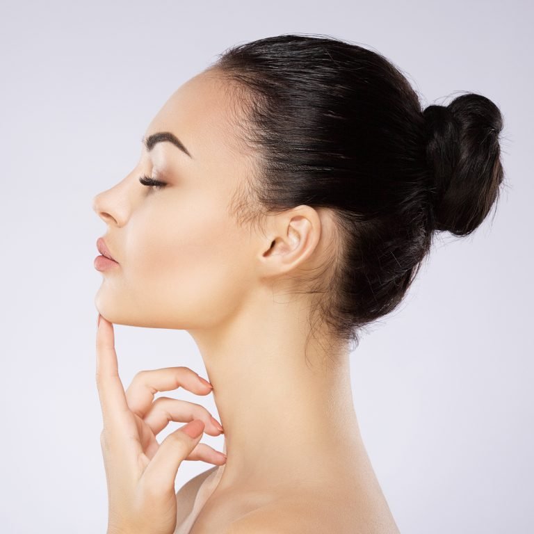 Cute girl with black hair fixed behind, thick eyebrows and naked shoulders holding hand near face at gray background, portrait, copy space.